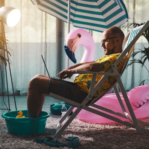 Man spending summer vacations at home alone, he is sitting on the deckchair in the living room and working with a laptop